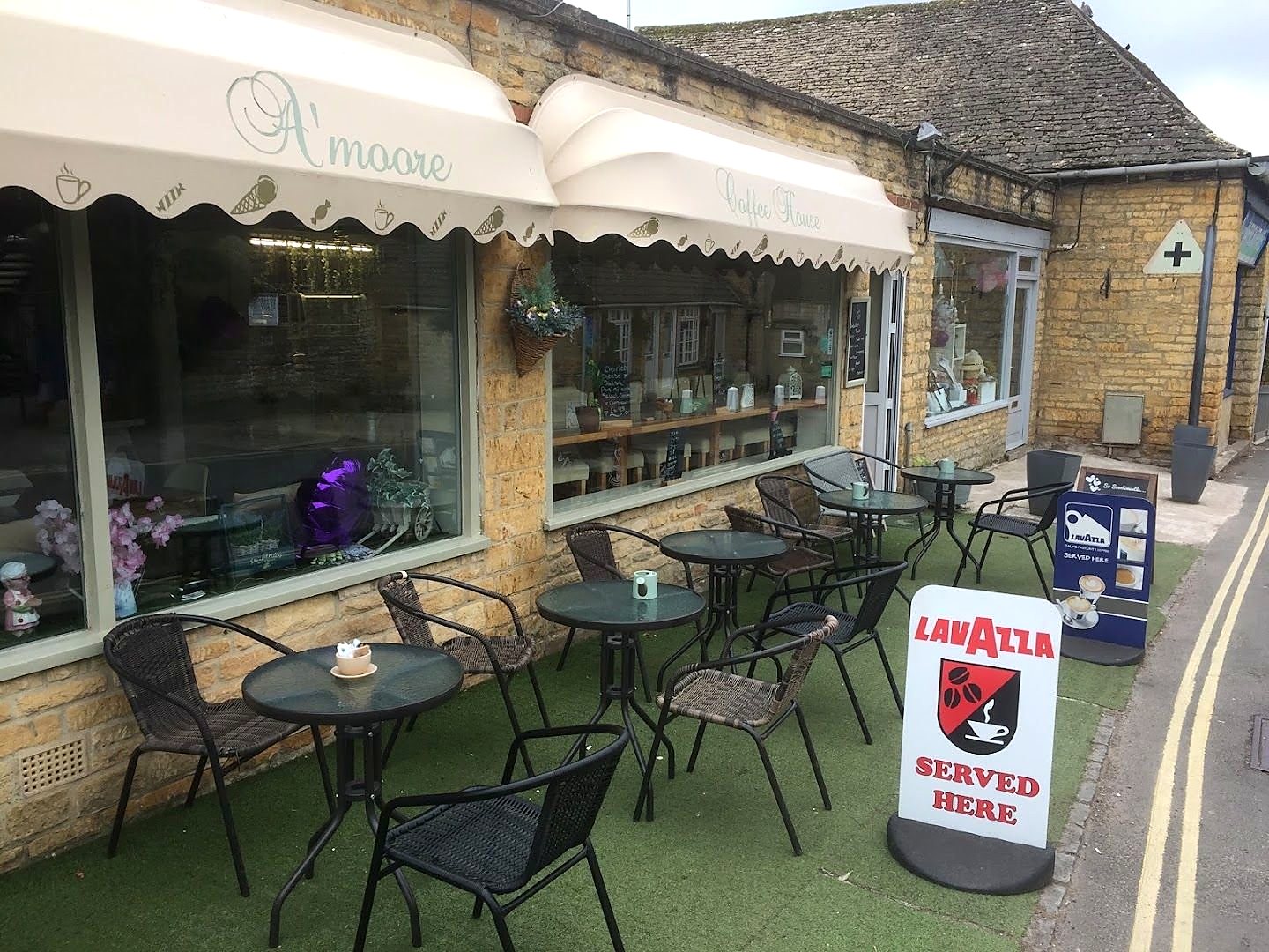 A'moore Coffee House: a Cotswold stone building with cream scalloped awnings, the name in script above the windows, black bistro tables on artificial grass and a Lavazza sign outside